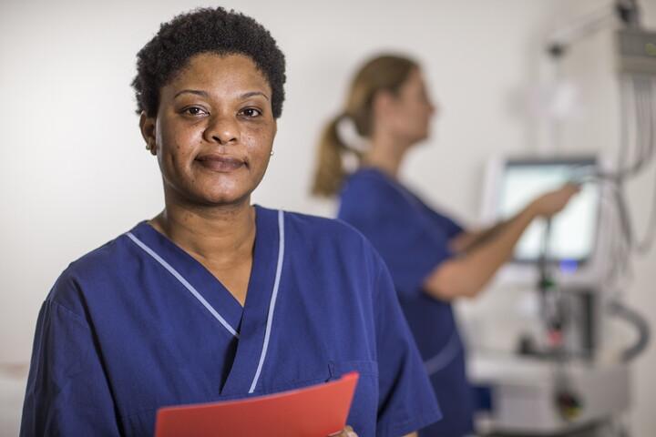 Kvinna i vårdkläder tittar in i kameran och håller en röd mapp i händerna.