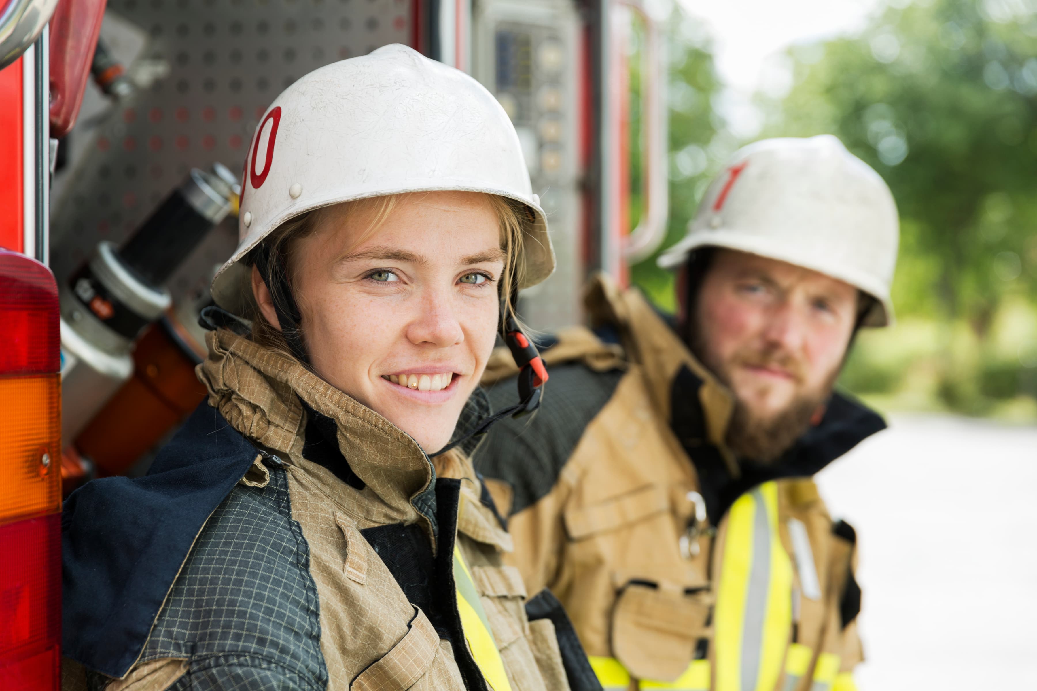 En kvinnlig och en manlig brandman i arbetskläder tittar in i kameran. Kvinnan ler.