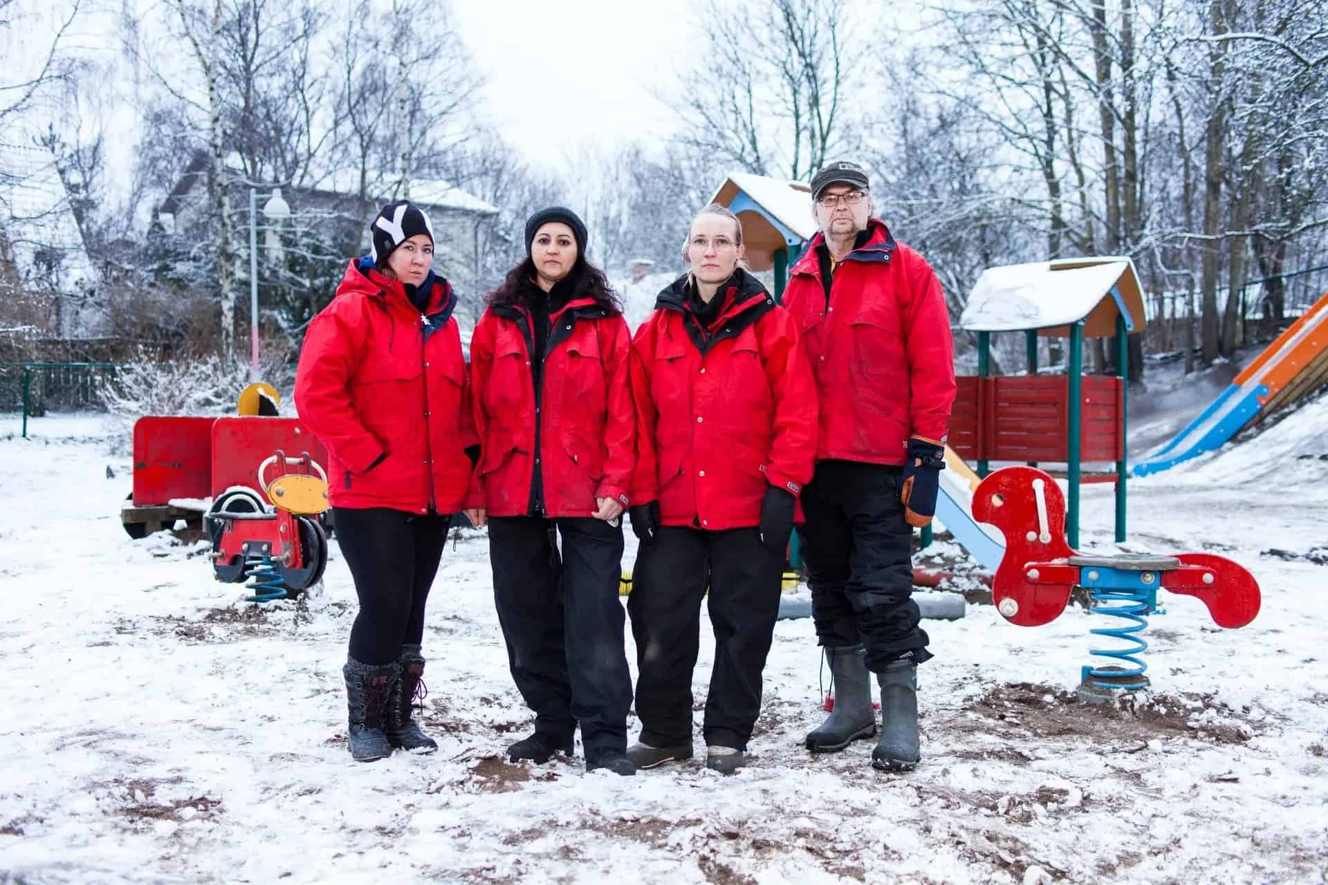 Fyra barnskötare i röda jackor står i en snöig lekpark.