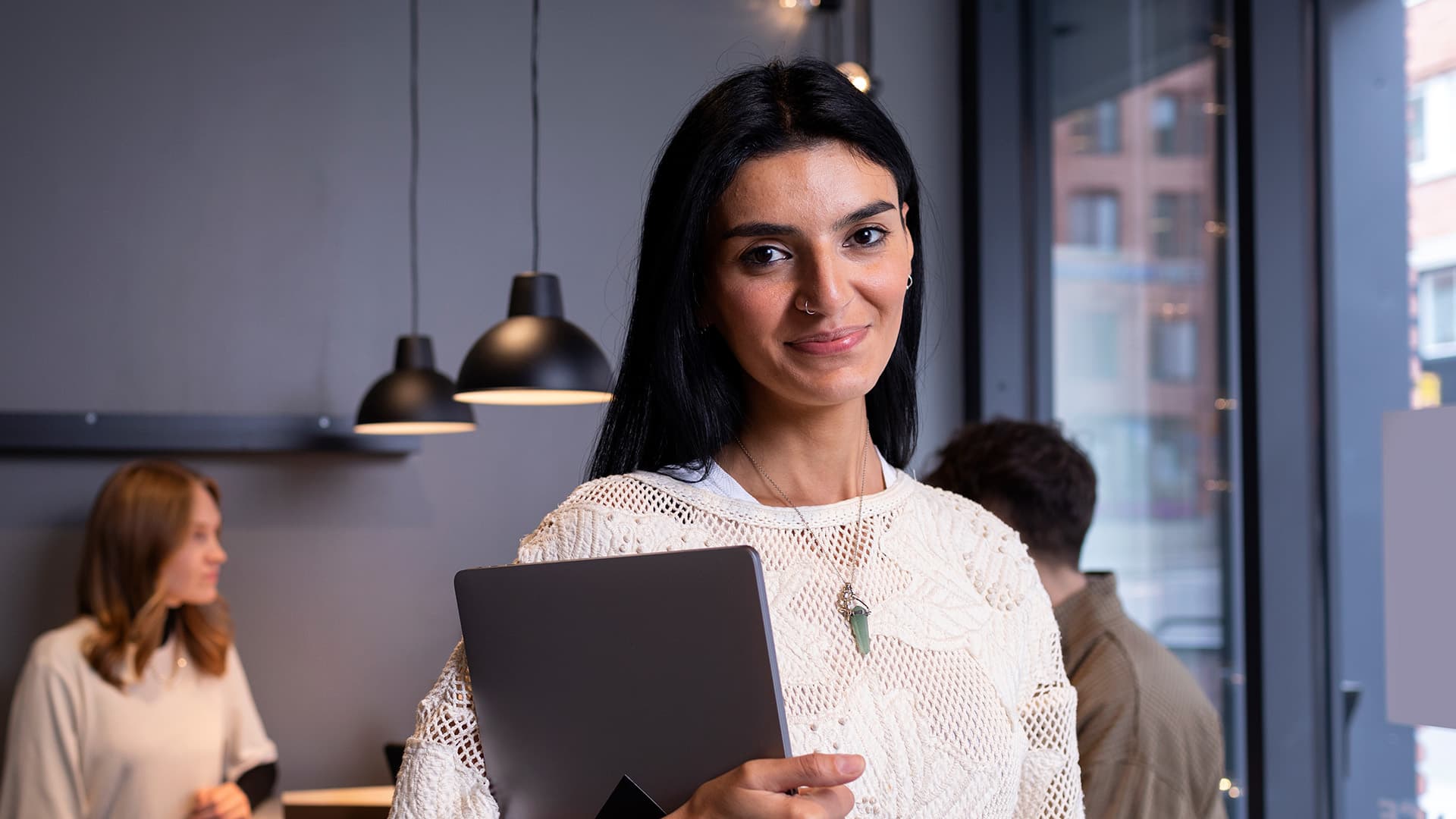 Kvinna med laptop och mobil som står framför kameran och ler.
