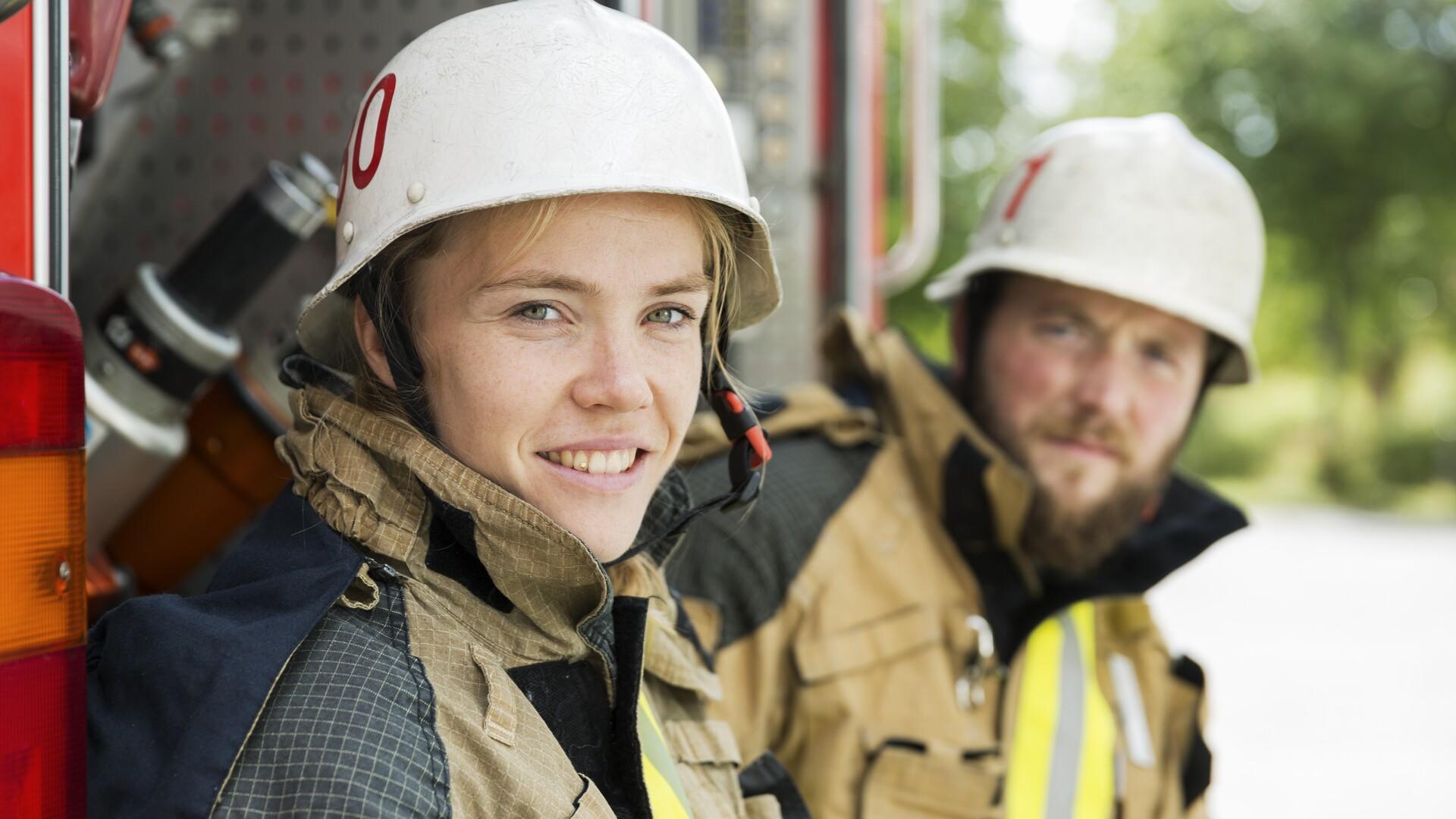 En kvinnlig och en manlig brandman i arbetskläder tittar in i kameran. Kvinnan ler.