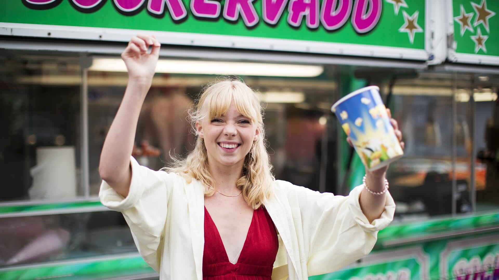 Glad tjej med händerna uppåt. Har popcornbehållare i högerhanden och står framför ett sockervaddstånd. Festival eller tivoli. 