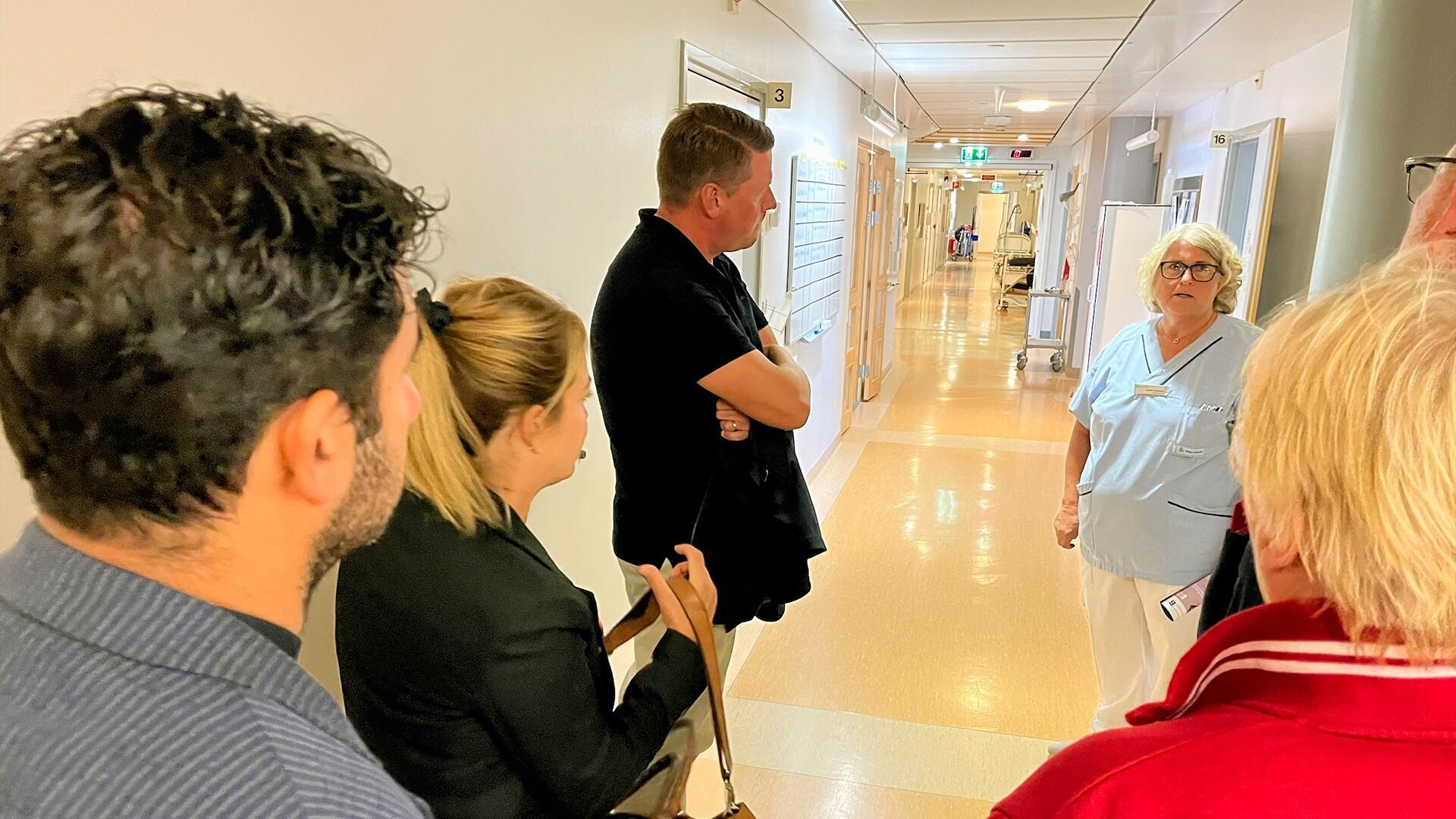 Personer i en sjukhuskorridor som lyssnar på en sjuksköterska eller chef.