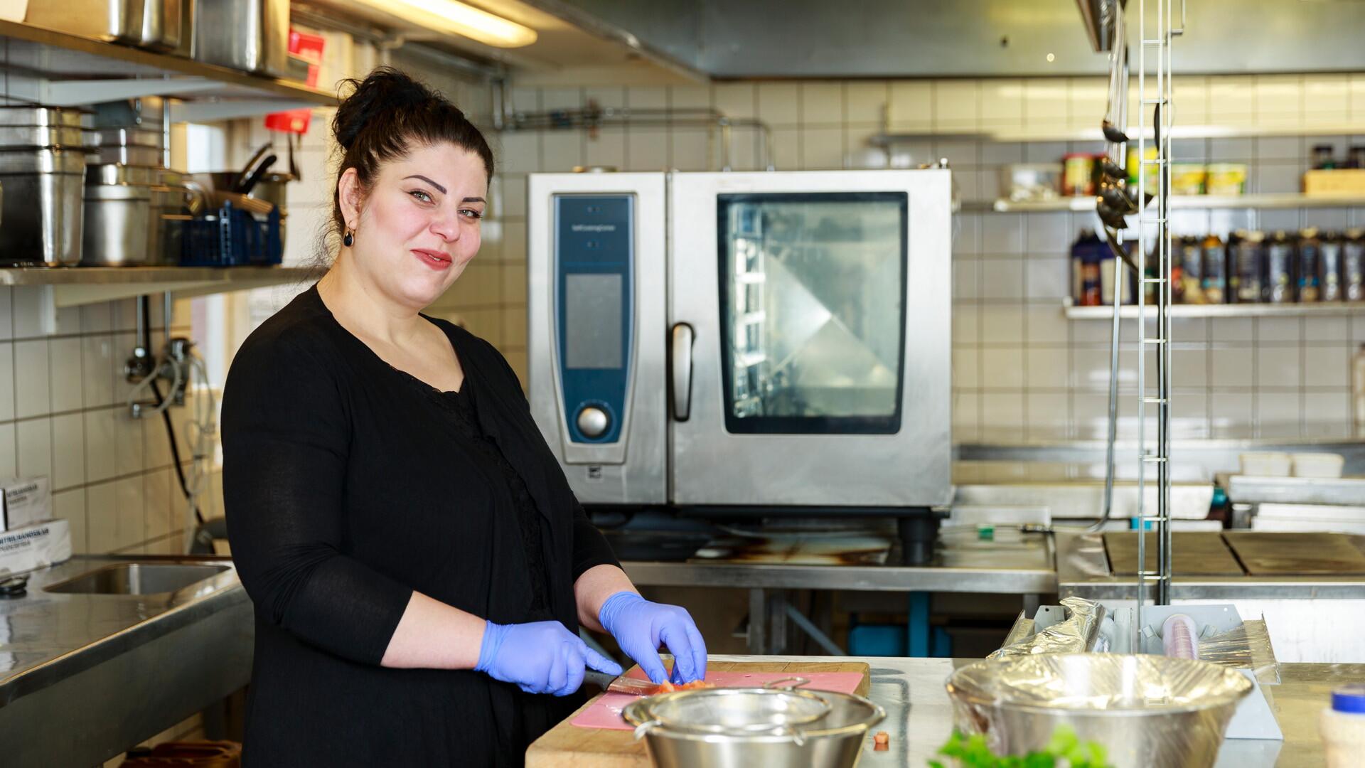 Kvinna står i ett restaurangkök. Hon har blå plasthandskar och skär grönsaker.