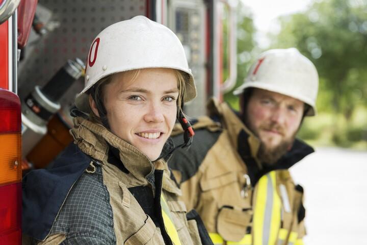 En kvinnlig och en manlig brandman i arbetskläder tittar in i kameran. Kvinnan ler.