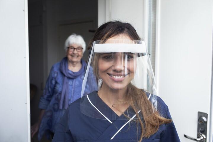 En sjuksköterska med visir ler mot kameran. Bakom henne står en äldre kvinna.