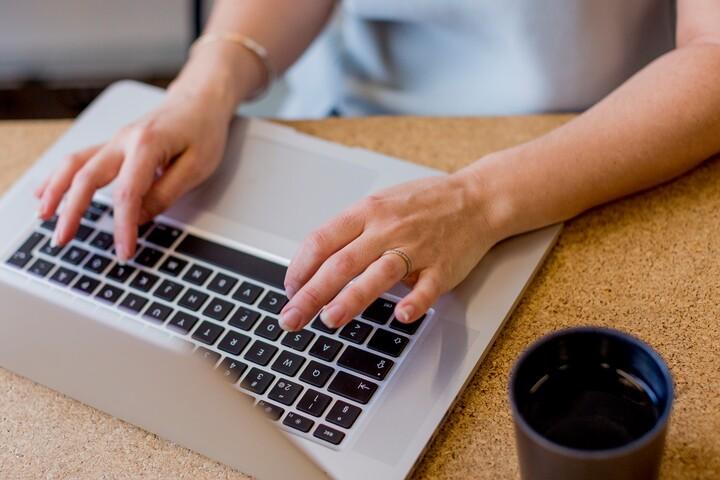 En bärbar dator står på ett bord. En kvinnas händer skriver på tangenterna. Bredvid datorn står en kaffekopp.
