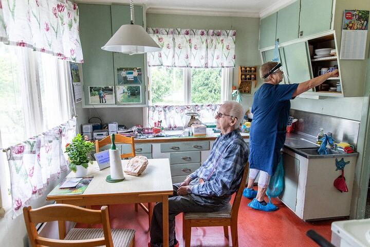Äldre man sitter vid köksbord och tittar ut medan kvinna ställer in saker i köksskåp bakom ryggen.
