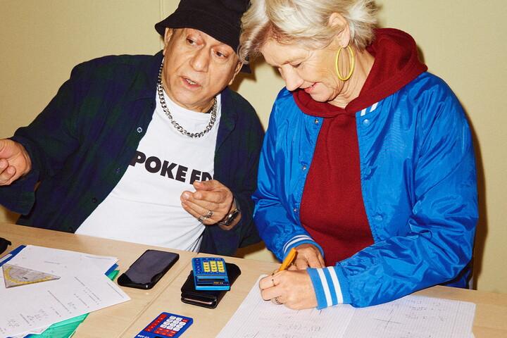 Två äldre personer med ungdomliga kläder sitter vid en skolbänk. På bordet ligger miniräknare och papper.
