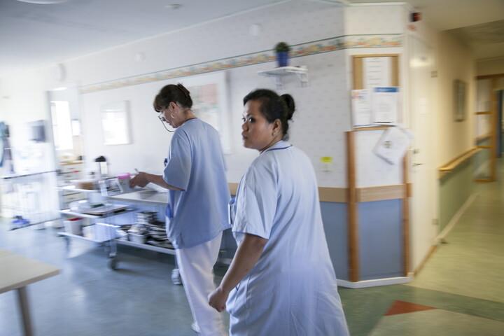 Två kvinnor med arbetskläder i vårdmiljö går runt hörnet i en korridor.